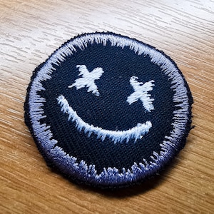 May include: A black embroidered patch with a white smiley face with X's for eyes. The patch has a light purple border.