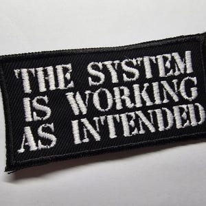 Op de afbeelding: Zwart rechthoekig embleem met wit geborduurde tekst: "THE SYSTEM IS WORKING AS INTENDED". Het embleem heeft een zwarte rand en is waarschijnlijk gemaakt van stof.