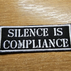 May include: Black and white embroidered patch with the text "Silence is Compliance" on a black background.