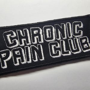 Pode incluir: Patch retangular preto com texto bordado branco que diz "CHRONIC PAIN CLUB". O patch tem uma borda preta e provavelmente foi projetado para ser costurado ou passado a ferro em roupas ou acessórios.
