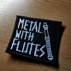 May include: Black embroidered patch with white text that reads "Metal with Flutes" and a white outline of a flute.