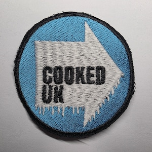 May include: Embroidered patch featuring a light blue circular background with a white arrow design. The text "COOKED UK" is in black. The arrow has an icy texture, and the patch has a black border.