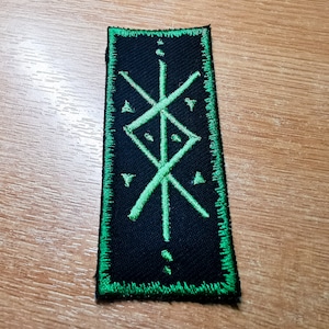 May include: Green embroidered patch with a black background featuring a symbol with a vertical line and two diagonal lines intersecting it. The symbol is surrounded by a green border.