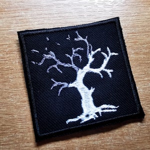 Puede incluir: Un parche de tela negro con una imagen bordada en blanco de un árbol estilizado con ramas desnudas. El árbol está centrado en el parche.