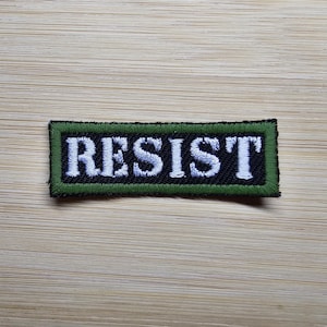 Könnte beinhalten: Gestickter Aufnäher mit dem Wort "RESIST" in weißen Großbuchstaben auf schwarzem Hintergrund. Der Aufnäher hat einen grünen Rand und ist rechteckig. Der Aufnäher liegt auf einer hellbraunen Holzoberfläche.