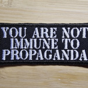 Può includere: Toppa rettangolare nera con testo ricamato bianco: "YOU ARE NOT IMMUNE TO PROPAGANDA". La toppa ha un bordo nero ed è su una superficie di legno marrone chiaro.