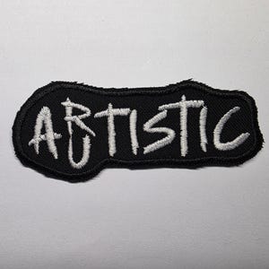 May include: Black embroidered patch with the word "ARTISTIC" in white, stylized lettering. The patch has a black border and is set against a white background. The patch is likely designed to be sewn onto clothing or accessories.