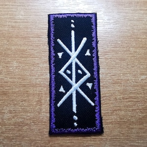 May include: A white embroidered symbol on a black patch with a purple border. The symbol is a stylized design with a vertical line and diagonal lines forming a diamond shape.
