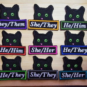 May include: Nine black cat head patches with different coloured backgrounds and text. The text on the patches reads "They/Them", "She/They", "He/Him", "He/Him", "She/Her", "He/They", "He/They", "She/They", and "She/Her".