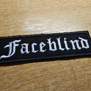 Puede incluir: Parche de tela negro con texto bordado en blanco que dice "Faceblind".