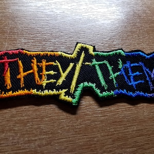 May include: An embroidered patch with the text "THEY/THEM" in black thread, with a rainbow-coloured border.