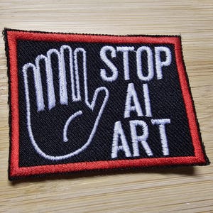 May include: A black patch with a red border, featuring a white hand in a stop gesture and the text "STOP AI ART".