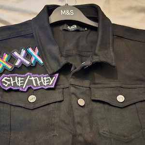 May include: A black denim vest with three patches sewn on the front. The patches are colorful and feature the text "XXX" and "SHE/THEY".