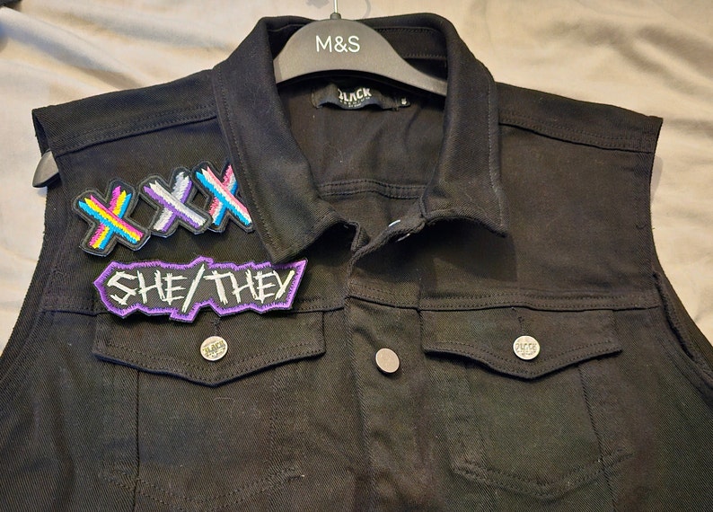 May include: A black denim vest with three patches sewn on the front. The patches are colorful and feature the text "XXX" and "SHE/THEY".