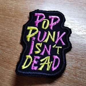 May include: Black embroidered patch with the text "Pop Punk Isn't Dead" in yellow and pink lettering.