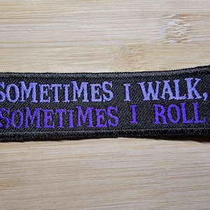 Puede incluir: Parche bordado rectangular negro con el texto "SOMETIMES I WALK, SOMETIMES I ROLL" en hilo morado. El parche est&aacute; sobre una superficie de madera marr&oacute;n claro.