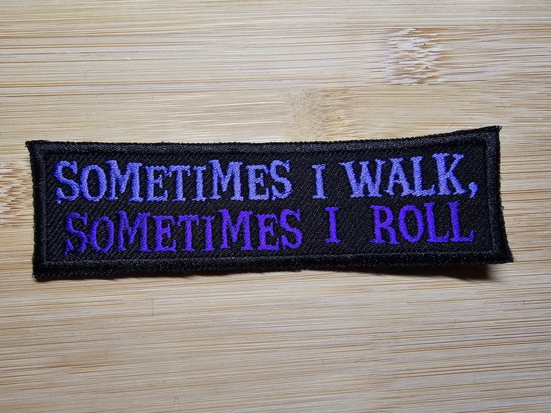 Puede incluir: Parche bordado rectangular negro con el texto "SOMETIMES I WALK, SOMETIMES I ROLL" en hilo morado. El parche est&aacute; sobre una superficie de madera marr&oacute;n claro.