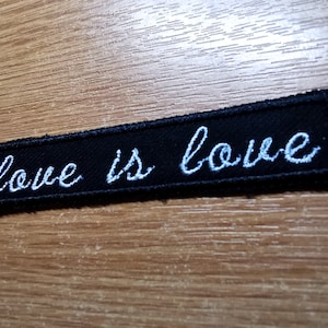 May include: Black embroidered patch with white cursive text that reads "love is love".