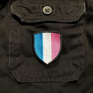 May include: A black fabric patch with a white shield shape. The shield has three vertical stripes: blue, white, and pink.