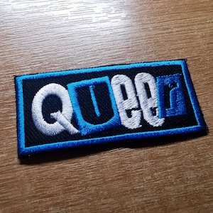 Pode incluir: Um remendo bordado azul e branco com a palavra "QUEEN" em letras brancas sobre um fundo preto.