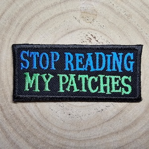 May include: Black embroidered patch with the text "STOP READING MY PATCHES" in blue and green lettering.