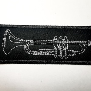 Könnte beinhalten: Ein schwarzer Stoff-Patch mit einer weißen gestickten Umrandung einer Trompete.