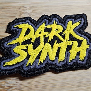 Puede incluir: Un parche bordado negro y amarillo con el texto "DARK SYNTH" en una fuente estilizada.