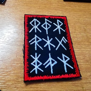 May include: A black fabric patch with a red border, featuring white embroidered runes. The runes are arranged in three rows, with three runes in each row.