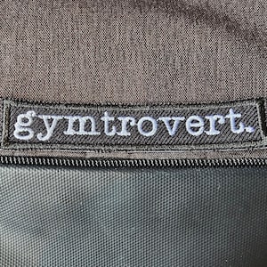 Puede incluir: Una bolsa de tela negra con un parche bordado en negro y blanco que dice "gymtrOvert."