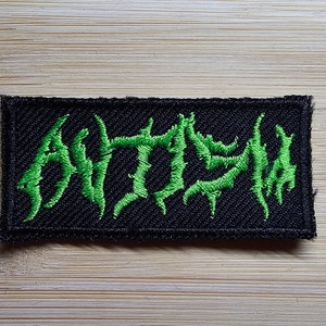 Könnte beinhalten: Grüner gestickter Aufnäher mit dem Wort "ANTHEM" in einer stilisierten Schriftart auf schwarzem Hintergrund.