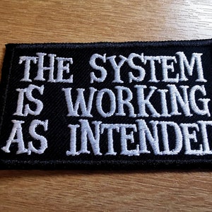 以下が含まれることがあります： 「THE SYSTEM IS WORKING AS INTENDED」と白い刺繍が施された黒い長方形のパッチ。木製の表面に配置されています。