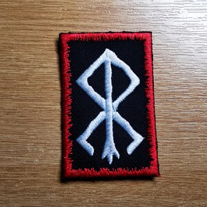 May include: A black embroidered patch with a white symbol, surrounded by a red border. The symbol is a stylized rune with a vertical line and four diagonal lines.