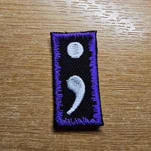 May include: A purple-bordered black fabric patch with a white semicolon embroidered on it.