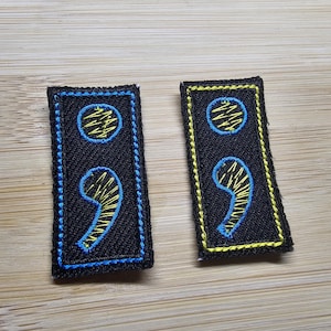 May include: Two black fabric patches with blue and yellow embroidered semicolon symbols. The patches have a blue and yellow border.