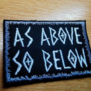 May include: Black rectangular patch with white embroidered text reading "AS ABOVE SO BELOW". The patch has a white stitched border and is set against a wooden surface. The text includes stylized symbols.