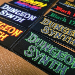 Pode incluir: Uma coleção de patches de tecido preto com o texto "Dungeon Synth" em várias cores, incluindo amarelo, verde, roxo e branco.