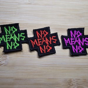 May include: Three black fabric patches in the shape of a cross with the words "NO MEANS NO" embroidered in green, red, and pink.