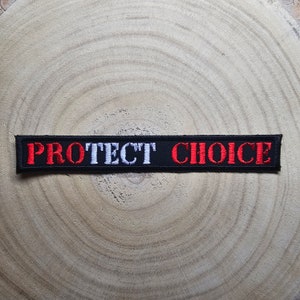 Op de afbeelding: Zwarte geborduurde patch met de woorden "PROTECT CHOICE" in rode en witte letters.