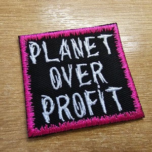 Puede incluir: Un parche de tela negro con costuras rosas alrededor de los bordes. El parche tiene texto blanco que dice "PLANET OVER PROFIT".
