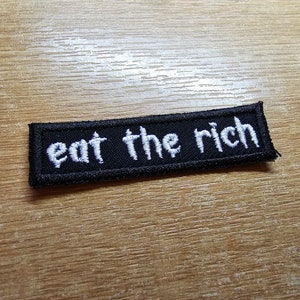 Puede incluir: Un parche rectangular negro con texto bordado en blanco que dice "eat the rich". El parche tiene un borde negro. El fondo es una superficie de madera.