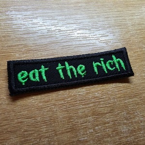 Peut inclure: Un écusson brodé noir et vert avec le texte "eat the rich" en vert.