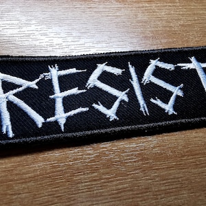 May include: White embroidered "RESIST" patch on a black fabric background.