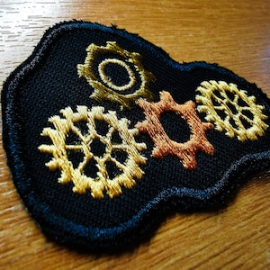 May include: Embroidered patch featuring a black background with three interlocking gears in shades of gold and copper. The patch has a dark blue border and is likely for clothing or accessories.