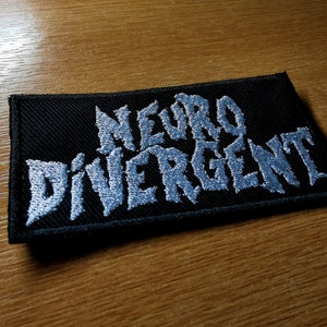 Puede incluir: Un parche de tela negro con texto bordado en blanco que dice "Neuro Divergent".