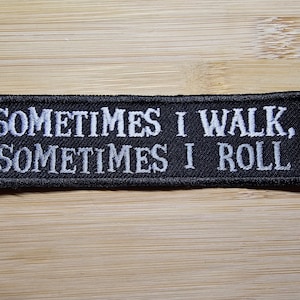 Puede incluir: Parche rectangular negro con texto bordado en blanco que dice: "SOMETIMES I WALK, SOMETIMES I ROLL". El parche est&aacute; sobre una superficie de madera marr&oacute;n claro. El parche tiene un borde negro.