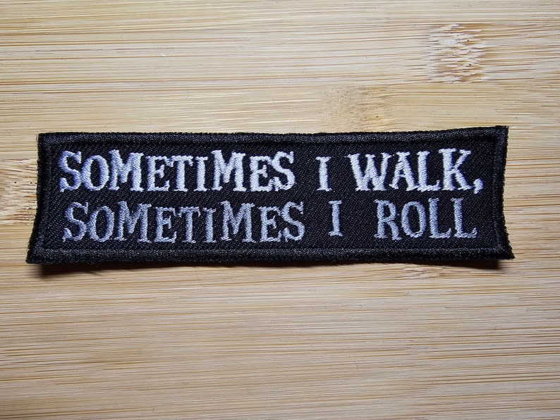 Puede incluir: Parche rectangular negro con texto bordado en blanco que dice: "SOMETIMES I WALK, SOMETIMES I ROLL". El parche est&aacute; sobre una superficie de madera marr&oacute;n claro. El parche tiene un borde negro.