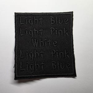 Puede incluir: Un parche cuadrado negro con texto bordado. El texto dice "LIGHT BLUE", "LIGHT PINK", "WHITE", "LIGHT PINK" y "LIGHT BLUE". El parche es probablemente para ropa o accesorios.