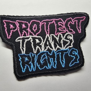 May include: Black embroidered patch with the words "PROTECT TRANS RIGHTS" in pink, white, and blue. The patch has a black border and a unique shape. The text is in a stylized font.