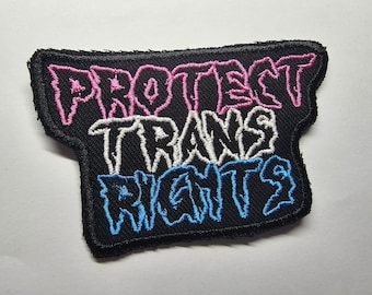 Puede incluir: Parche bordado negro con las palabras "PROTECT TRANS RIGHTS" en rosa, blanco y azul. El parche tiene un borde negro y una forma única. El texto está en una fuente estilizada.