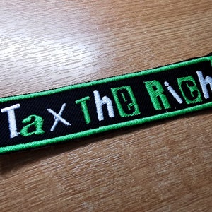 Op de afbeelding: Een zwart en groen geborduurd patch met de tekst "Tax The Rich" in wit.